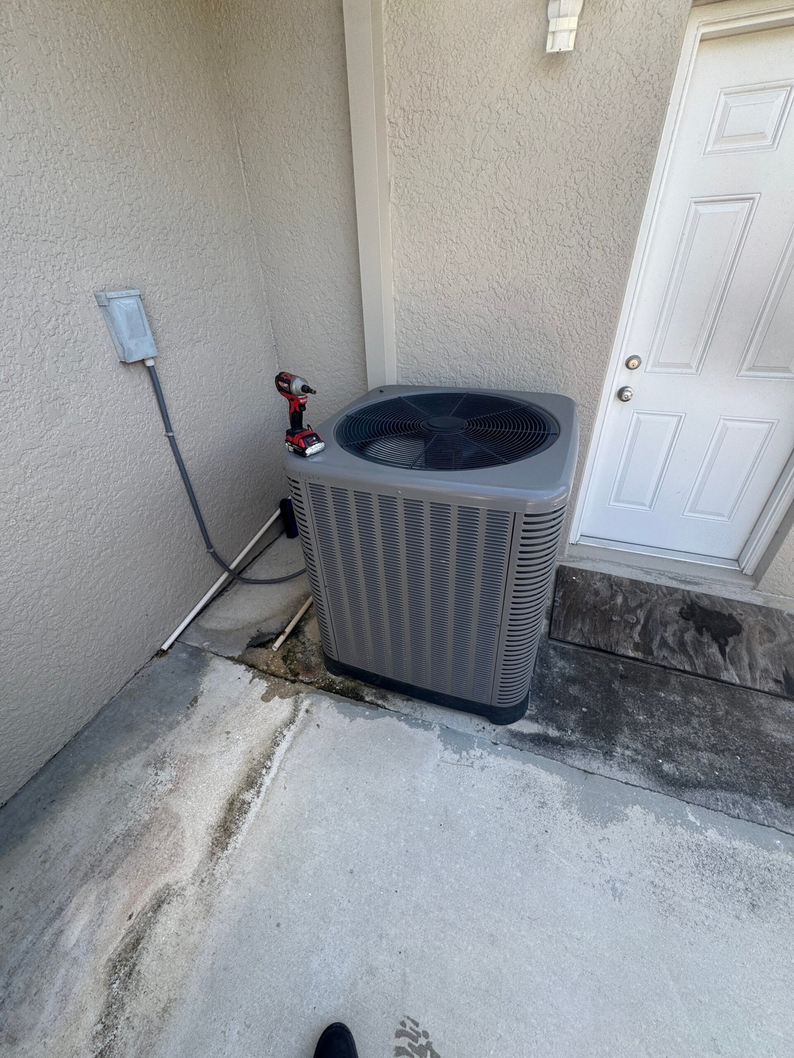 A modern, grey outdoor AC unit positioned between a textured stucco wall and a white service door in Cape Coral, Florida, with a power drill visible on top during a tune-up by technician Ryan.